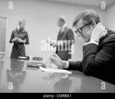 1970s WORRIED BUSINESSMAN SEATED CONFERENCE TABLE TWO PEOPLE IN BACKGROUND Stock Photo