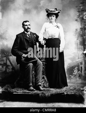 1890s 1900 TURN OF THE CENTURY PORTRAIT SERIOUS COUPLE HOLDING HANDS TOGETHER IN RUSTIC STUDIO SETTING Stock Photo