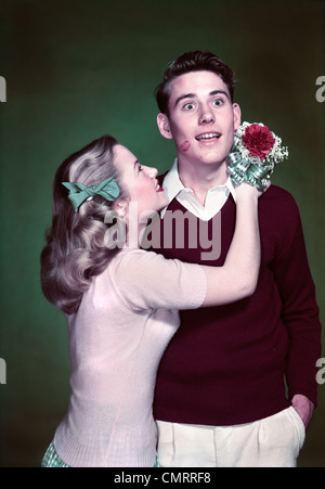 1940s 1950s TEEN COUPLE GIRL HOLDING NOSEGAY BOUQUET HUGGING SURPRISED BOY LIPSTICK KISS ON CHEEK Stock Photo