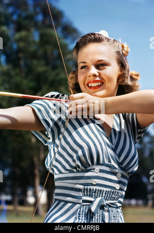 1940s 1950s SMILING TEEN GIRL ARCHER WEARING BLUE AND WHITE STRIPED DRESS SHOOTING AIMING BOW AND ARROW Stock Photo
