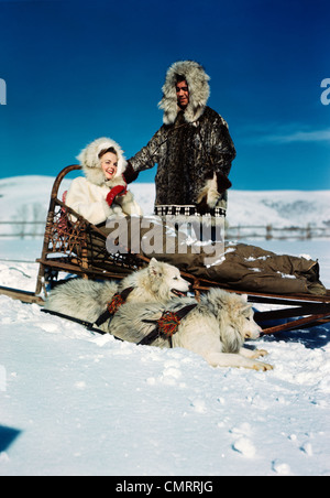 1940s 1950s COUPLE WEARING FUR LINED PARKAS WOMAN SITTING IN DOG SLED NEXT TO TWO DOGS HUSKIES Stock Photo