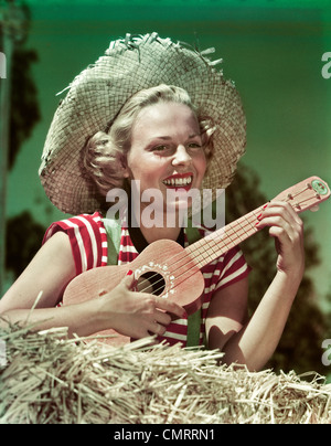 1940s 1950s SMILING BLOND WOMAN WEARING STRAW HAT SITTING ON HAY PLAYING UKULELE Stock Photo