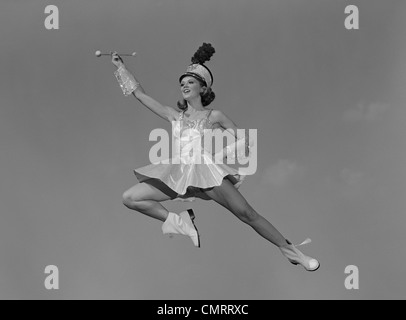 1960s WOMAN MAJORETTE HOLDING BATON WEARING SHORT SKIRT UNIFORM JUMPING INTO THE AIR Stock Photo
