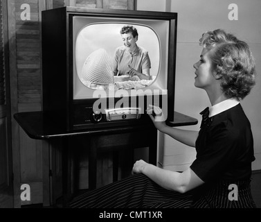 1950s BLONDE WOMAN TURNING DIAL ON TV SET WATCHING PROGRAM ABOUT BABY CHILDCARE Stock Photo