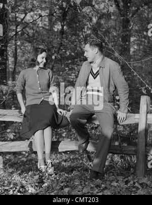 1940s YOUNG COUPLE SITTING ON FENCE AUTUMN SMILING AT ONE ANOTHER Stock Photo