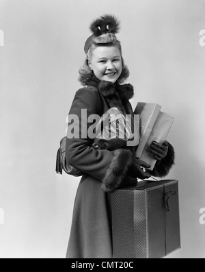 1940s 1950s SMILING YOUNG BLOND WOMAN WEARING HAT FUR TRIMMED COAT CARRYING PACKAGES LOOKING AT CAMERA Stock Photo