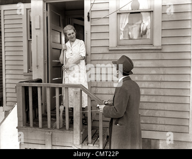 1940s 1930s MAN DOOR-TO-DOOR SALESMAN TALKING WOMAN TO HOUSEWIFE AT BACK DOOR Stock Photo