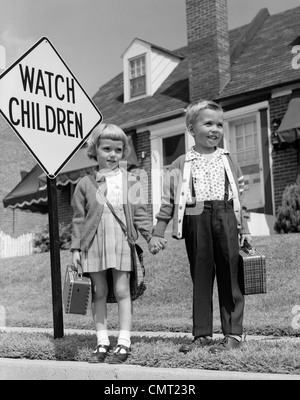 1960s BOY & GIRL DRESSED UP FOR SCHOOL CARRYING PLAID LUNCHBOXES HOLDING HANDS STANDING BY WATCH CHILDREN SIGN Stock Photo