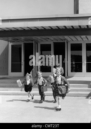 1950s 1960s FOUR YOUNG CHILDREN WITH BAGS AND LUNCH BOXES RUNNING FROM SCHOOL ENTRANCE SMILING Stock Photo