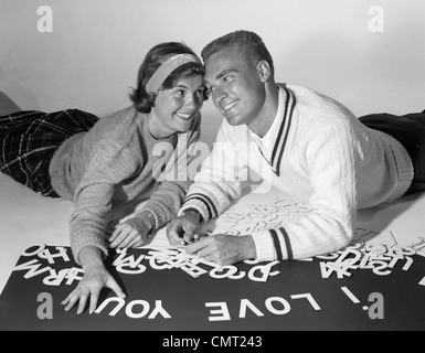 1960s SMILING COLLEGIATE COUPLE LYING ON STOMACHS WITH HEADS PRESSED TOGETHER SPELLING OUT I LOVE YOU WITH CUT-OUT LETTERS Stock Photo