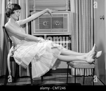 1960s SIDE VIEW OF WOMAN IN GINGHAM DRESS PROPPING LEGS UP ON STOOL PUTTING HAND ON TOP OF AIR CONDITIONER Stock Photo