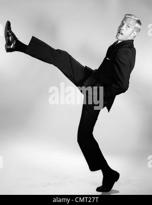 1960s SIDE VIEW OF MAN IN SUIT WITH HANDS IN POCKETS KICKING A FOOT AND LEG OUT HIGH IN THE AIR LOOKING AT CAMERA Stock Photo