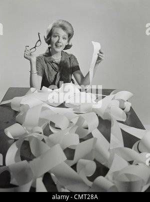 1960s WOMAN HOLDING EYEGLASSES AND END OF EXTREMELY LONG TAPE FROM OFFICE MACHINE Stock Photo