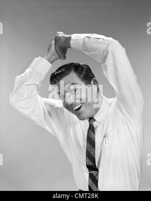 1960s BUSINESSMAN IN SHIRTSLEEVES CLASPING HANDS TOGETHER OVERHEAD SMILING SYMBOLIC SUCCESS WINNING STUDIO INDOOR Stock Photo