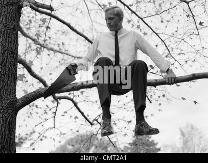 1960s BUSINESSMAN IN SHIRT AND TIE SITTING ON AND SAWING OFF TREE LIMB Stock Photo