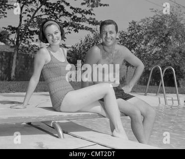 1960s MAN WOMAN COUPLE SITTING ON DIVING BOARD ON SIDE OF SWIMMING POOL Stock Photo