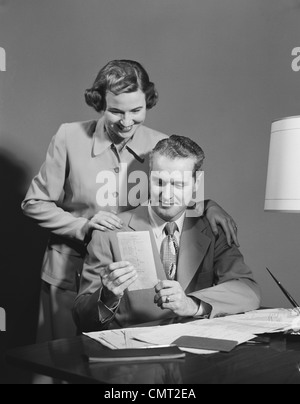 1950s COUPLE MAN SEATED AT DESK HOLDING PAPER WOMAN STANDING LOOKING OVER HIS SHOULDER SMILING HAPPY Stock Photo