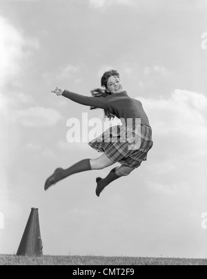 1960s CHEERLEADER IN SWEATER PLAID SKIRT & KNEE SOCKS LEAPING HIGH IN AIR LOOKING AT CAMERA Stock Photo