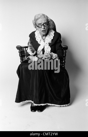 1970s WOMAN GRANDMOTHER IN OLD FASHIONED DRESS SITTING ROCKING CHAIR KNITTING SURPRISED FACIAL EXPRESSION LOOKING AT CAMERA Stock Photo
