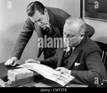 1930s 1940s TWO SALESMEN LOOKING AT SALES BROCHURE SITTING AT A DESK WITH CLIENT CARD FILE Stock Photo