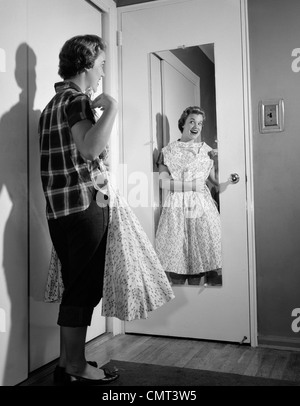 1950s TEEN GIRL IN JEANS HOLDING FORMAL DRESS UP TO CHEST LOOKING IN MIRROR ON BACK OF BEDROOM DOOR Stock Photo