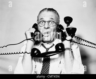 1950s 1960s PORTRAIT OF FRAZZLED BUSINESSMAN TRYING TO ANSWER FOUR BLACK TELEPHONES PHONES AT ONCE Stock Photo