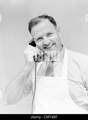 1930s 1940s MAN WEARING WHITE APRON SMILING TALKING ON TELEPHONE STORE CLERK BUTCHER BAKER GROCER SERVICE LOOKING AT CAMERA Stock Photo