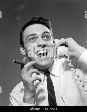 1950s 1960s EXCITED MAN TALKING ON TELEPHONE GRINNING SMOKING CIGAR INDOOR Stock Photo