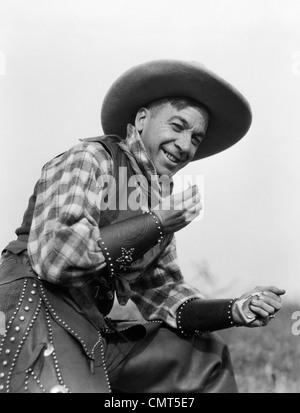 1920s SMILING MAN IN COWBOY HAT VEST CUFFS AND CHAPS ROLLING CIGARETTE Stock Photo