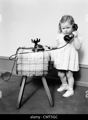 1930s 1940s UNHAPPY FROWNING LITTLE GIRL TALKING ON OLD BLACK ROTARY DIAL TELEPHONE Stock Photo