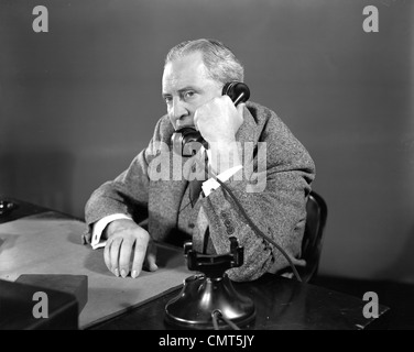 1930s 1940s SUSPICIOUS BUSINESSMAN TALKING ON TELEPHONE SITTING AT DESK Stock Photo