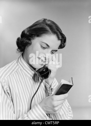 1950s YOUNG WOMAN TALKING ON TELEPHONE READING ADDRESS FROM LITTLE BLACK BOOK Stock Photo