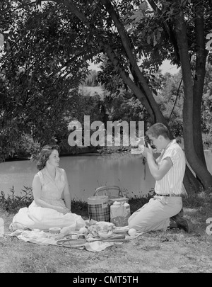 1950s TEENAGE COUPLE PICNIC BOY TAKING PHOTOGRAPH OF GIRL OUTDOORS Stock Photo