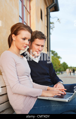 Man and woman using computer while leaning on house wall Stock Photo