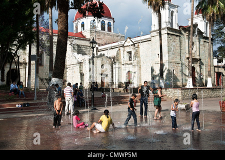 parents & children frolic in water play feature in Juarez Llano park presided over by Our Lady of Guadalupe church Oaxaca Stock Photo