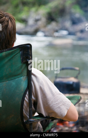 Man relaxing in chair next to river on camping expedition.  Wooroonooran National Park, Queensland, Australia Stock Photo