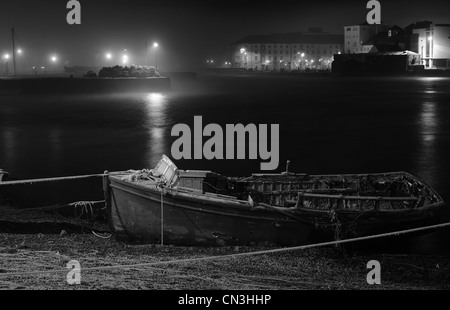 A monochrome shot of an old dilapidated boat tied up in Galway Bay on a misty night with dim lamp lights brightening the horizon Stock Photo