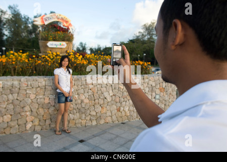 A young man photographs a friend on his mobile phone on the promenade by the entrance to Songkhla Lake from the sea, Songkhla, Thailand Stock Photo