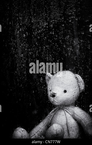 Sad Teddy bear looking through a window covered in rain drops. Monochrome. Still life Stock Photo