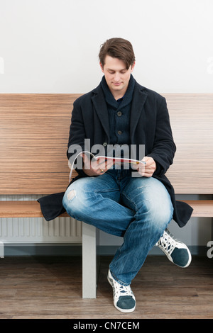 Young man sitting in the waiting room of a hospital, reading a magazine to kill the time Stock Photo
