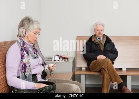 Elderly woman, sitting on a bench in the waiting room of a hospital with a portable music player in her hands. Stock Photo