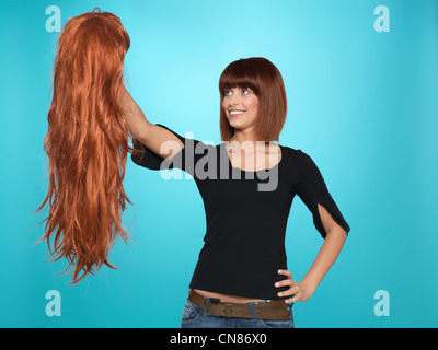 beautiful, young woman admiring a long, red hair wig she is holding, on blue background Stock Photo