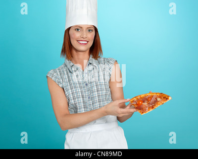 portrait of beautiful young woman chef, holding a pizza slice in one of her hands, on blue background Stock Photo