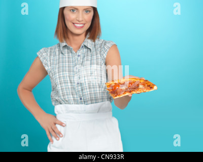 portrait of beautiful young woman chef, holding a pizza slice in one of her hands, on blue background Stock Photo