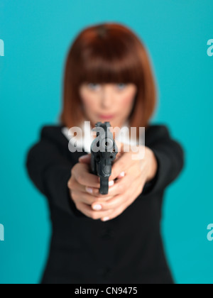 portrait of an agressive, beautiful, young businesswoman, pointing a gun in front of her, on blue background Stock Photo