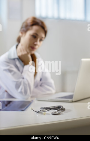 closeup of stethoscope on desk, with mid adult female doctor working on laptop in background Stock Photo