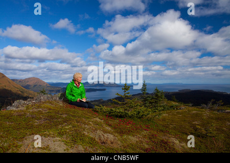 Mature woman relaxes and writes in journal on Pillar Mountain with Monashka Bay in background, Kodiak Island, Alaska, Autumn Stock Photo
