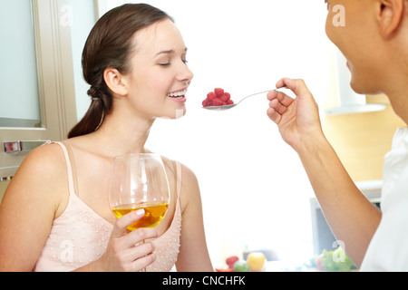 Image of happy woman with glass of wine looking at spoonful of raspberries in her husband hand Stock Photo