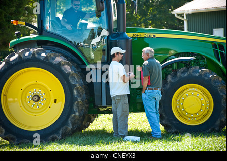 Two men standing next to a large tractor and one man riding inside of the tractor Stock Photo