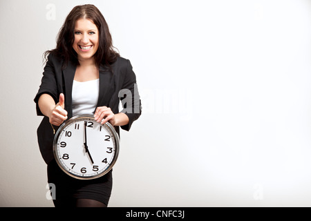 Portrait of a female employee giving a thumbs up sign while holding a clock, isolated on gray background Stock Photo